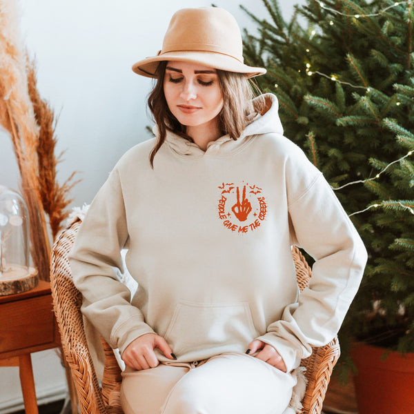 A woman wearing a beige hat and a white hoodie with an orange graphic is sitting in a wicker chair, with a potted plant and a glass object in the background.