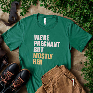 A green t-shirt with text printed on it, along with some pants and a pair of shoes, all placed on a surface with greenery.