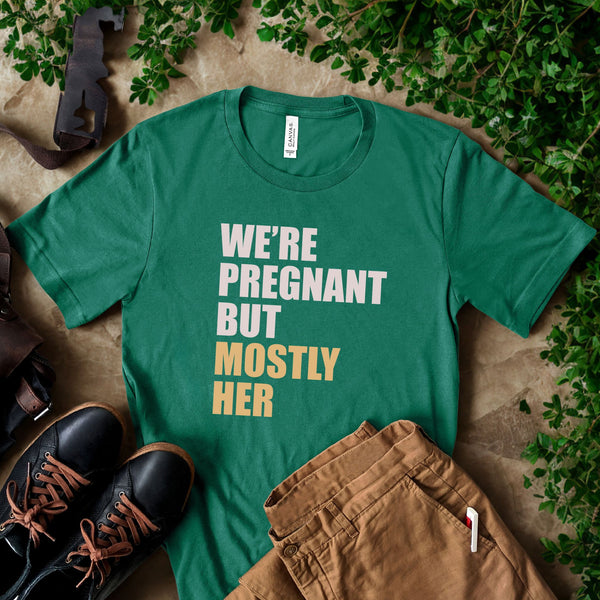 A green t-shirt with text printed on it, along with some pants and a pair of shoes, all placed on a surface with greenery.