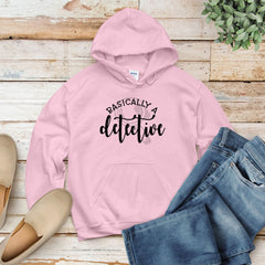 A pink hoodie with the text "BASICALLY A DETECTIVE" printed on it, along with a pair of jeans and a pair of shoes, all laid out on a wooden surface.