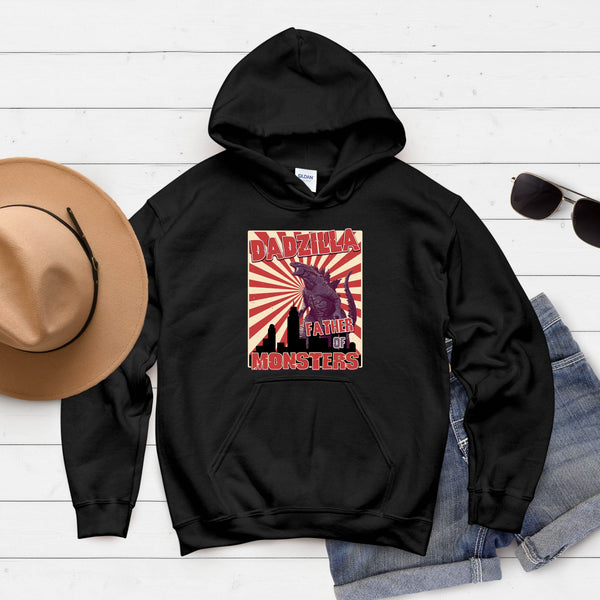 A black hoodie with a graphic design featuring a large purple dinosaur and the text "DADZILA FATHER OF MONSTERS" printed on it. The hoodie is placed on a white surface, along with a hat and sunglasses.