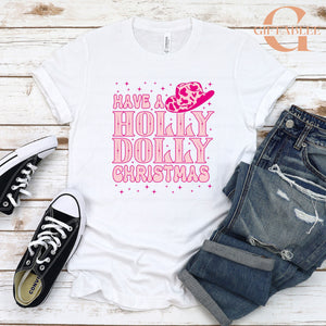 A white t-shirt with a pink and purple design, featuring the text "Have a Holly Dolly Christmas" and a small pink cowboy hat icon. The t-shirt is placed on a wooden surface, along with a pair of black and white sneakers and a pair of blue jeans.