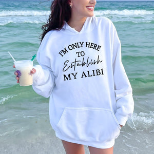 A woman wearing a white hoodie with the text "I'M ONLY HERE TO ESTABLISH MY ALIBI" standing on a beach with the ocean in the background.