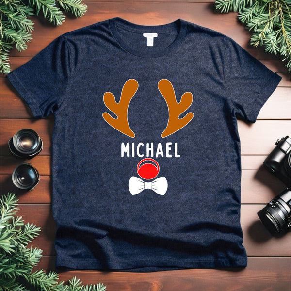 A navy blue t-shirt with a graphic design of a reindeer and the name "MICHAEL" printed on it, along with a red nose and a white bowtie. The t-shirt is placed on a wooden surface, surrounded by pine branches.